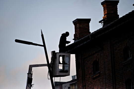 Fototapeta Człowiek na wysięgniku naprawiający oświetlenie na dachu zabytkowego budynku z cegły czerwonej , kominy i widoczna latarnia uliczna.