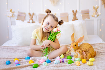 Obraz na płótnie Canvas easter, a little girl with painted colored eggs and a rabbit holding spring flowers tulips at home in a bright room preparing for the holiday smiling and having fun