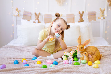 easter, a little girl with painted colored eggs, tulips and a rabbit at home in a bright room is preparing for the holiday smiling, having fun and playing with eggs closing her eyes