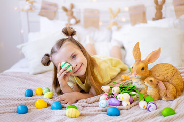 easter, a little girl with painted colored eggs, tulips and a rabbit at home in a bright room is preparing for the holiday smiling, having fun and playing with eggs closing her eyes