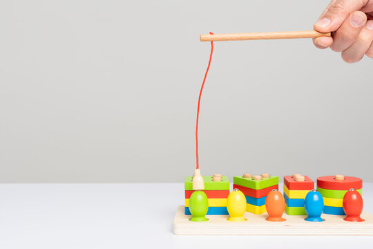 A hand with a toy fishing rod with a magnet shows how to use a wooden sorter toy with colored puzzles and figures to simulate fishing. Children's educational game