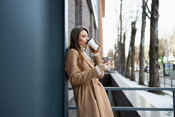 Side view of a young woman in a brown coat drinking hot coffee and using a mobile phone outdoor