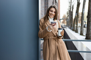 Pretty brunette girl in brown coat coat with coffee cup using smartphone outdoor