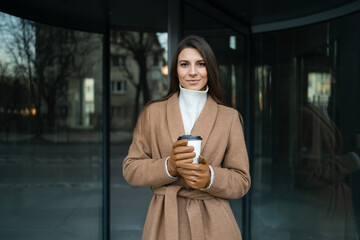 Woman in a brown coat with a cup of coffee on the background of the glass doors of office building