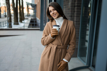 Cheerful woman in brown coat with coffee cup outdoors