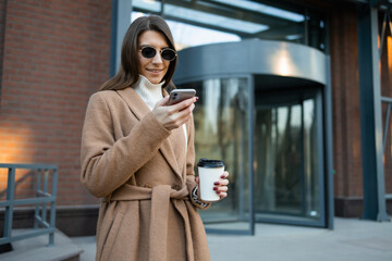 Stylish woman in brown coat and glasses with coffee cup using smartphone outdoors