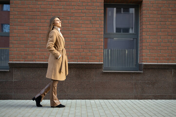 Side view of beautiful woman in brown coat and leather pants near brick building