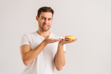 Portrait of dissatisfied young man with disgust pointing to bad burger on white isolated background. Studio shot of handsome bearded male holding in hands unhealthy delicious hamburger.