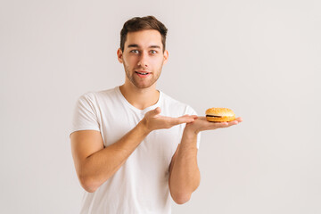 Portrait of handsome young man pointing to tasty burger on white isolated background, looking at camera. Studio shot of happy bearded male holding in hands unhealthy delicious hamburger.