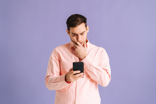 Portrait of frustrated young man holding phone with doubtful and skeptical expression on pink isolated background. Studio shot of handsome male thinking and reading online message using smartphone.