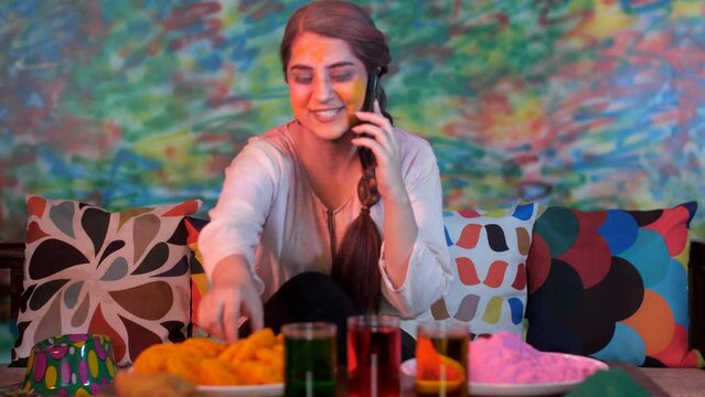 A beautiful young girl wearing a white Kurti talking on her mobile on Holi - leisure time  festive season. A pretty female with yellow Gulal on her face enjoys eating Imarti while relaxing in her l...