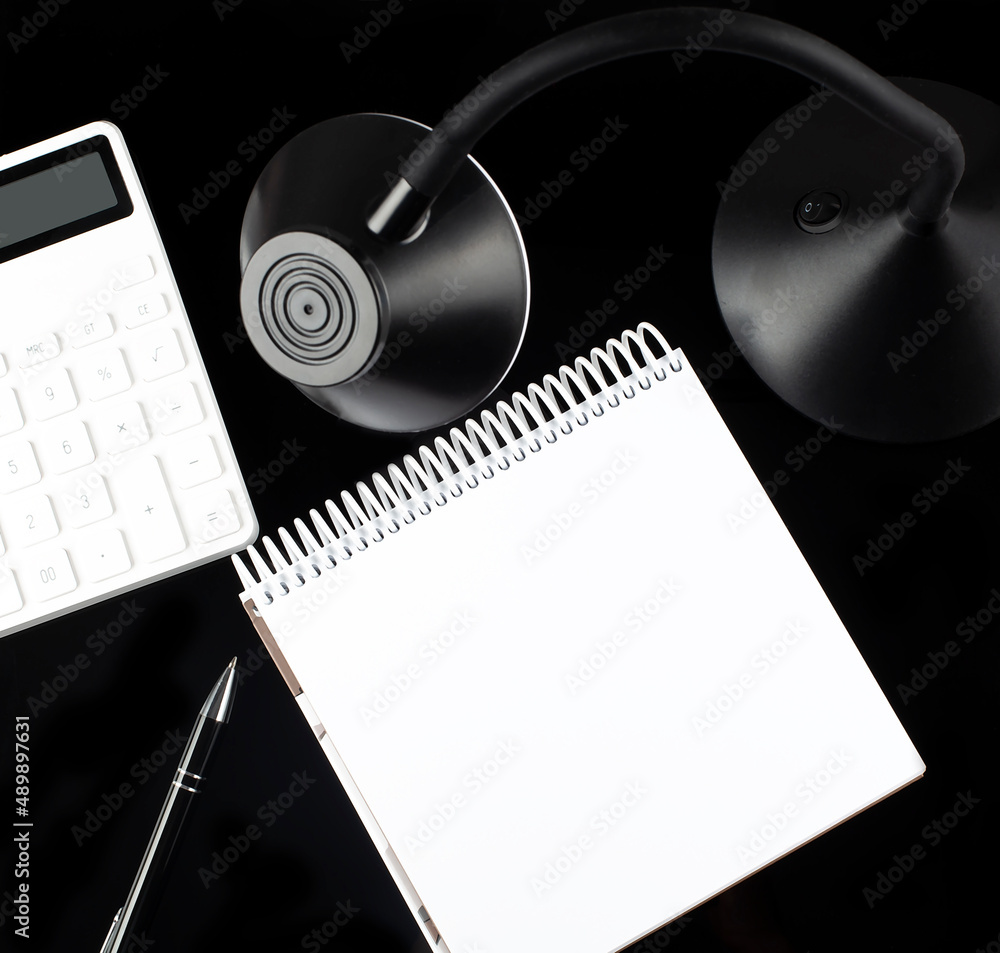 Poster opened paper notebook with a pen, calculator and table lamp on black background.