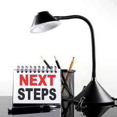 NEXT STEPS text on notebook with pen and table lamp on the black background