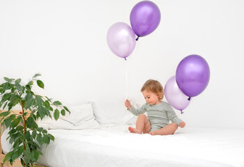 Fototapeta na wymiar Cute funny caucasian blonde baby girl,toddler of 1,2 year old playing on bed with big purlple balloons of very peri trendy color on white background in modern scandinavian interior at home.Copy space