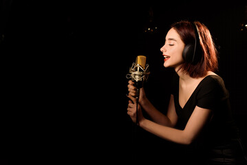 Young woman with headphones is recording a song in an audio recording studio on black background.