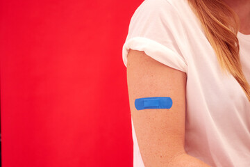 Woman With Blue Sticking Plaster On Arm After Injection Or Vaccination Against Red Background