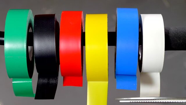 Closeup of a finger and thumb pulling a piece of yellow sticky electrical tape from one of the line of multi color rolls, then cutting a piece off with a pair of scissors.