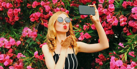 Obraz na płótnie Canvas Beautiful woman taking selfie picture by phone blowing her red lips sending sweet air kiss on pink flowers roses background