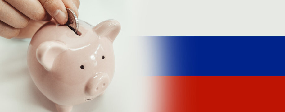 Close up of woman putting a coin inside piggy bank as investment against background of Russian national flag.