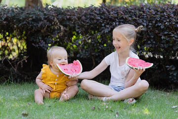 Cute little caucasian girl and her baby sister eating watermelon outdoor at hot summer day. Kids siblings spending time together and having fun during summer vacations