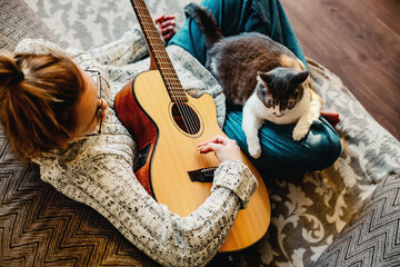 Girl musician sits on couch and plays guitar. Cat sits on mistress's lap. Playing musical...