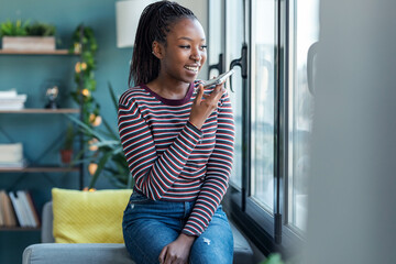 Smiling young african american woman sending audio message with mobile phone while sitting on sofa...