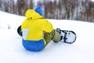 A young man in a yellow overall sits on a mountain top with a snowboard against the background of a forest. The snowboarder is preparing to go down the snowy slope. End of the ski season. © Евгений Панов
