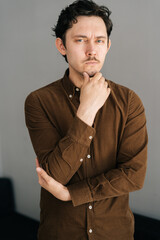 Vertical portrait of serious young man with little moustache holding hand on chin and thoughtful looking at camera, thinking over problem, standing on background of grey wall.