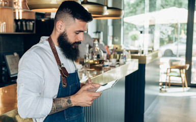 Handsome Barista Using Digital Tablet in a Cafe. Serious waiter with a beard standing at bar counter and reading online order or watching something on his tablet while working in the restaurant.