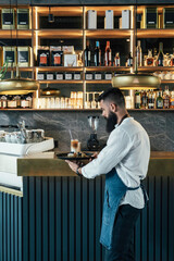 Handsome Waiter Carries Iced Coffee and Dessert on a Serving Tray. 
Professional barista standing at bar counter and preparing an order of latte coffee and slice of cake in a coffee shop.