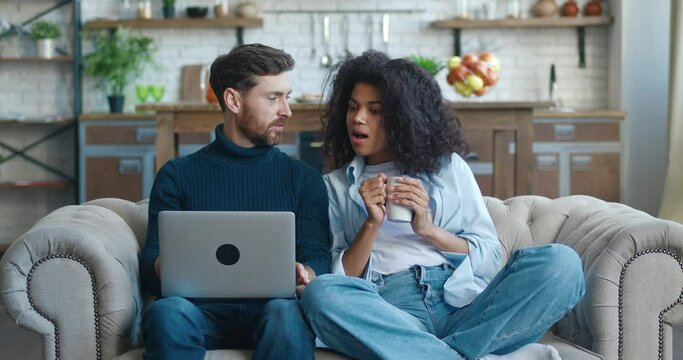 Happy interracial young couple using laptop for ecommerce doing online shopping together watching video movie sitting on sofa, smiling caucasian man and african woman looking at computer at home