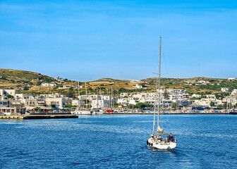 Fototapeta na wymiar Sailboat going to port of Greek island. Yachts and fishing boats in harbor at sunny summer day. Whitewashed houses on green hills.
