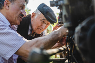 Obraz na płótnie Canvas Two caucasian senior men father and son pensioner at the farm repairing the tractor machine in sunny day family bonding generation cooperation and togetherness concept real people