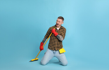 Handsome young man with floor brush singing on light blue background
