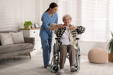 Senior woman in wheelchair doing physical exercise and young caregiver helping her indoors. Home...