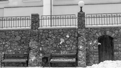 Black and white image of wooden benches with snow and ice. The seating area is directly in front of a stone wall.