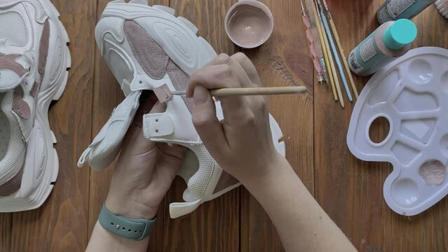 Female's hands dip brush in brown paint and apply it to sneaker. Design of sports shoes using acrylic. Palette, tools and tubes on wooden table. Hobbies, manual work and leisure at home.