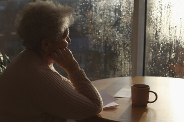 Elderly woman with drink looking out of window indoors on rainy day, space for text. Loneliness...