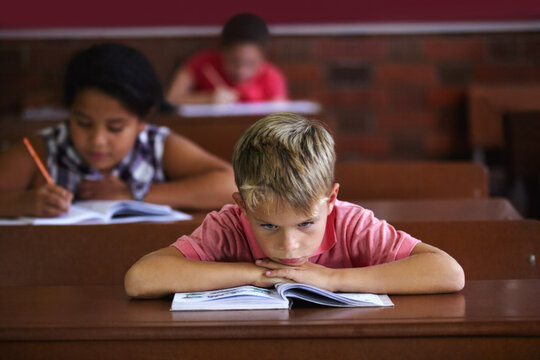 School Sucks. A Young Boy Resting His Head On His Arms As He Sits In A Classroom Looking Bored.