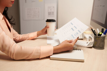 Pensive female UI designer drinking cup of coffee and looking at printed mockup of mobile application