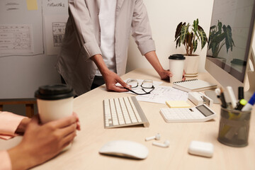 Cropped image of UI designer putting cup of coffee on table of coworker with drawing of wireframes...