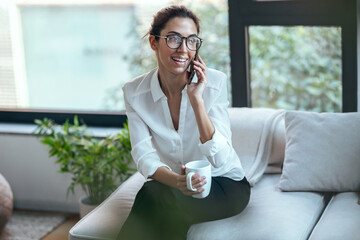 Pretty young woman using mobile phone while drinking cup of coffee on sofa at home.