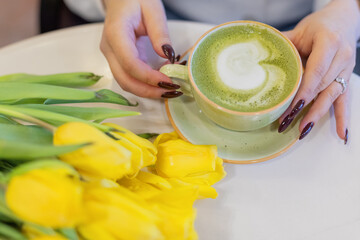 a girl holds a cup of matcha green tea in her hands. flowers on the table in front of her. View from above. Top view. A cup of coffee. Hot breakfast drink. Hands close up