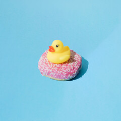Small yellow rubber duck toy on donut with pink glaze and small white crumbs. Blue pastel background. Minimal design. Copy space.