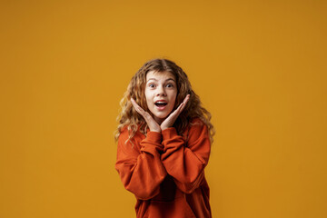 Portrait of a happy teenage girl with curly hair in a hoodie, smiling and rejoicing. A child, emotionally excited with delight, laughs with an open smile, standing on a yellow background in the studio