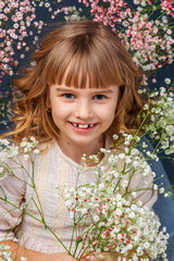Naklejka premium Close-up portrait of cute smiling little girl with curly hair and white dress. child is holding a bouquet of flowers, gypsophiles, standing on gray background in studio.