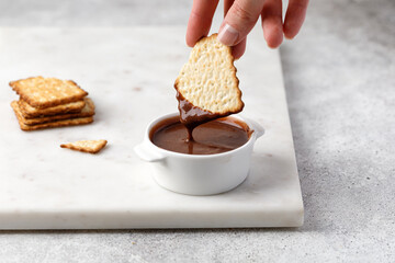 Crackers or cookies in chocolate paste in white sauce pan. A hand holds a cookie.