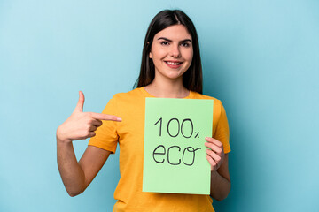 Young caucasian woman holding 100% eco placard isolated on blue background person pointing by hand to a shirt copy space, proud and confident