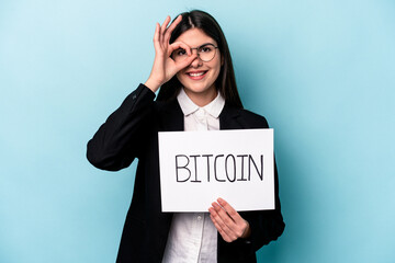 Young caucasian business woman holding a bitcoin placard isolated on blue background excited keeping ok gesture on eye.