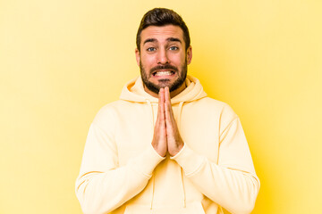 Young caucasian man isolated on yellow background holding hands in pray near mouth, feels confident.